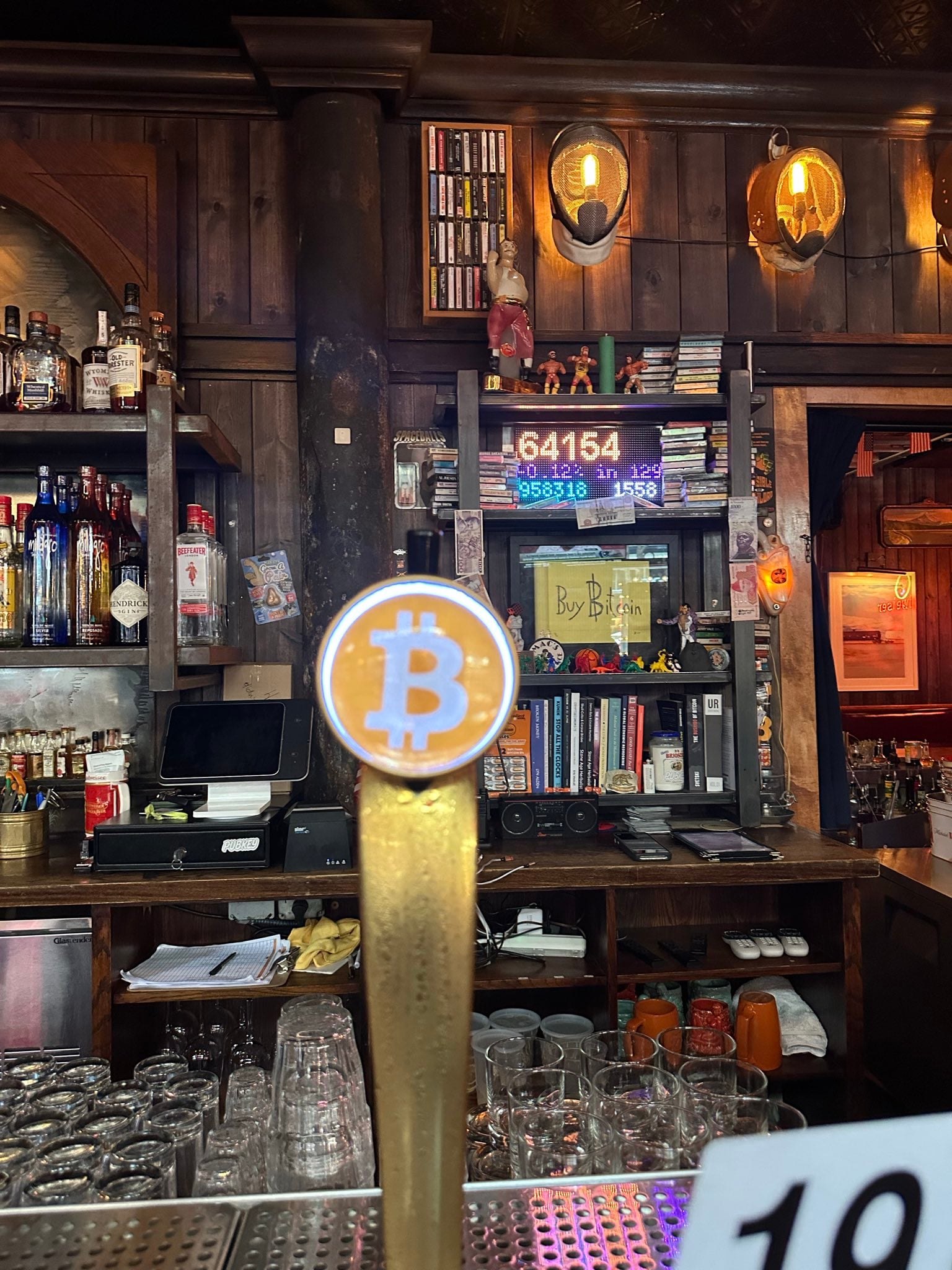 A beer tap handle capped with a glowing orange Bitcoin logo, with the back bar and the blocktron behind it.