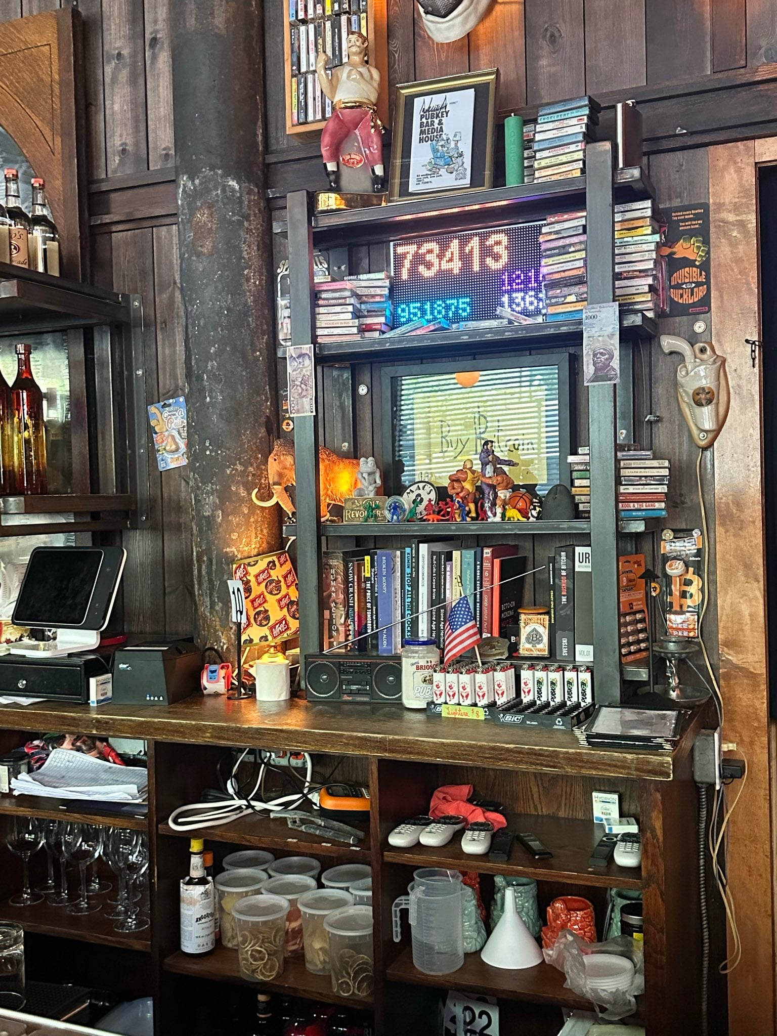 The blocktron LED board behind the bar, glowing with the current block height above shelves of cassettes, books, and bar oddities.